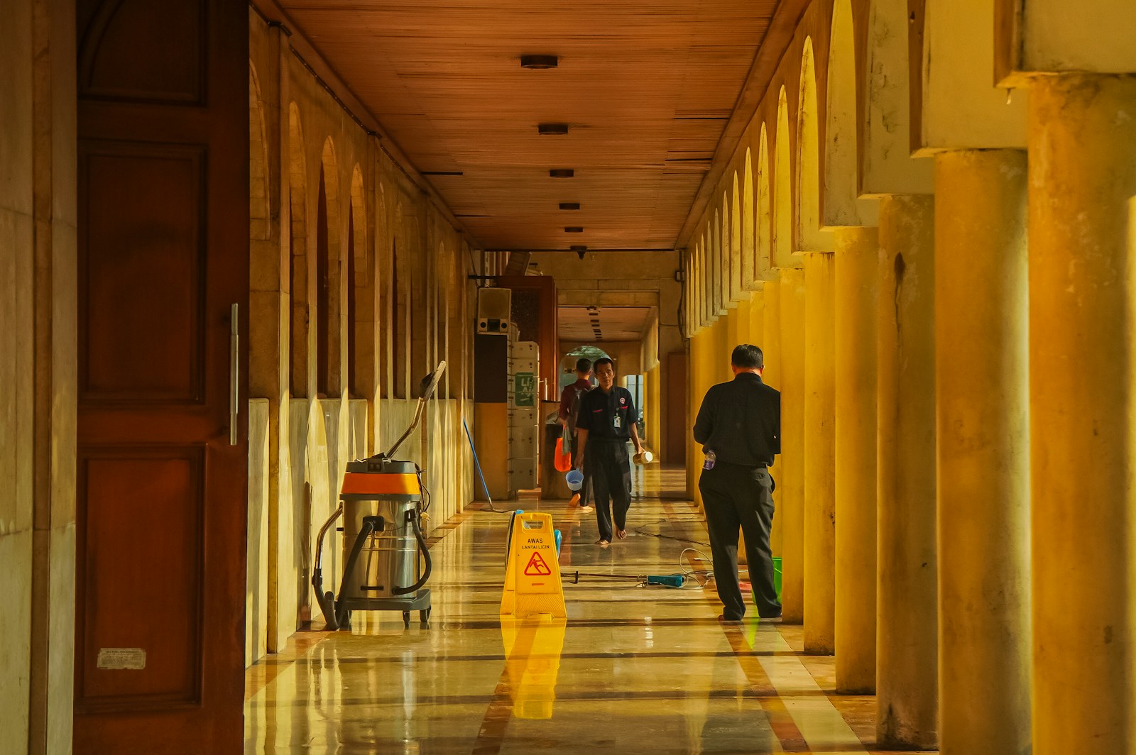 People walking down a long, sunlit hallway with columns