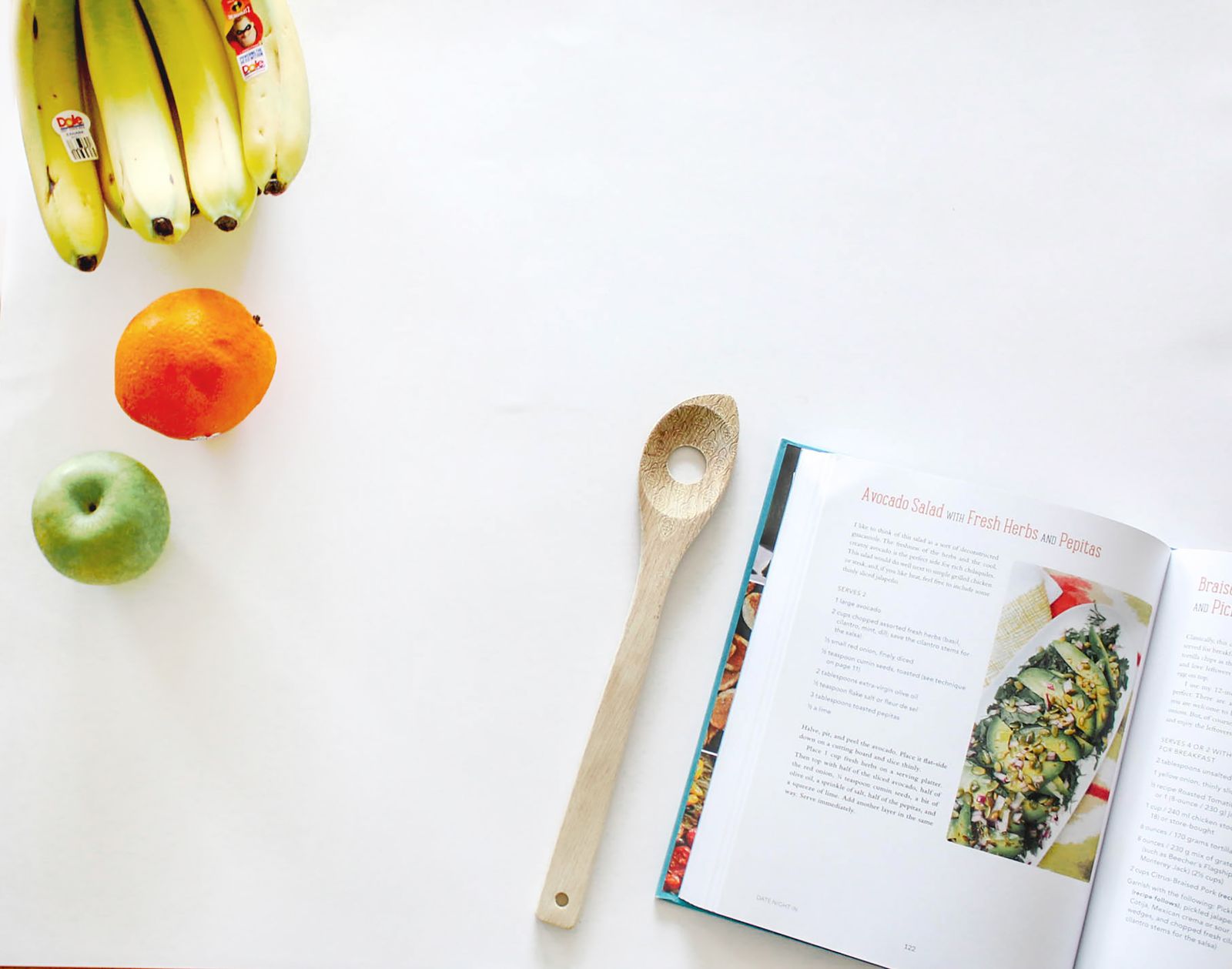 An open cookbook beside fruit and a wooden spoon