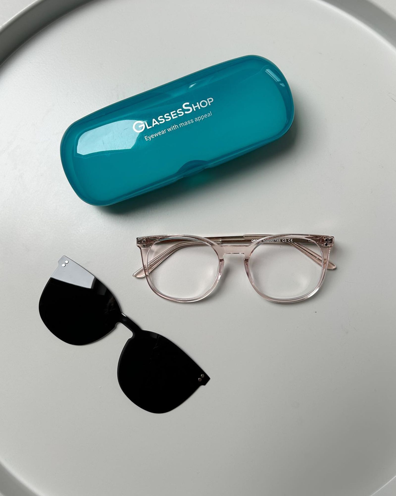 Eyeglasses, clip-on shades, and a teal glasses case on a white surface