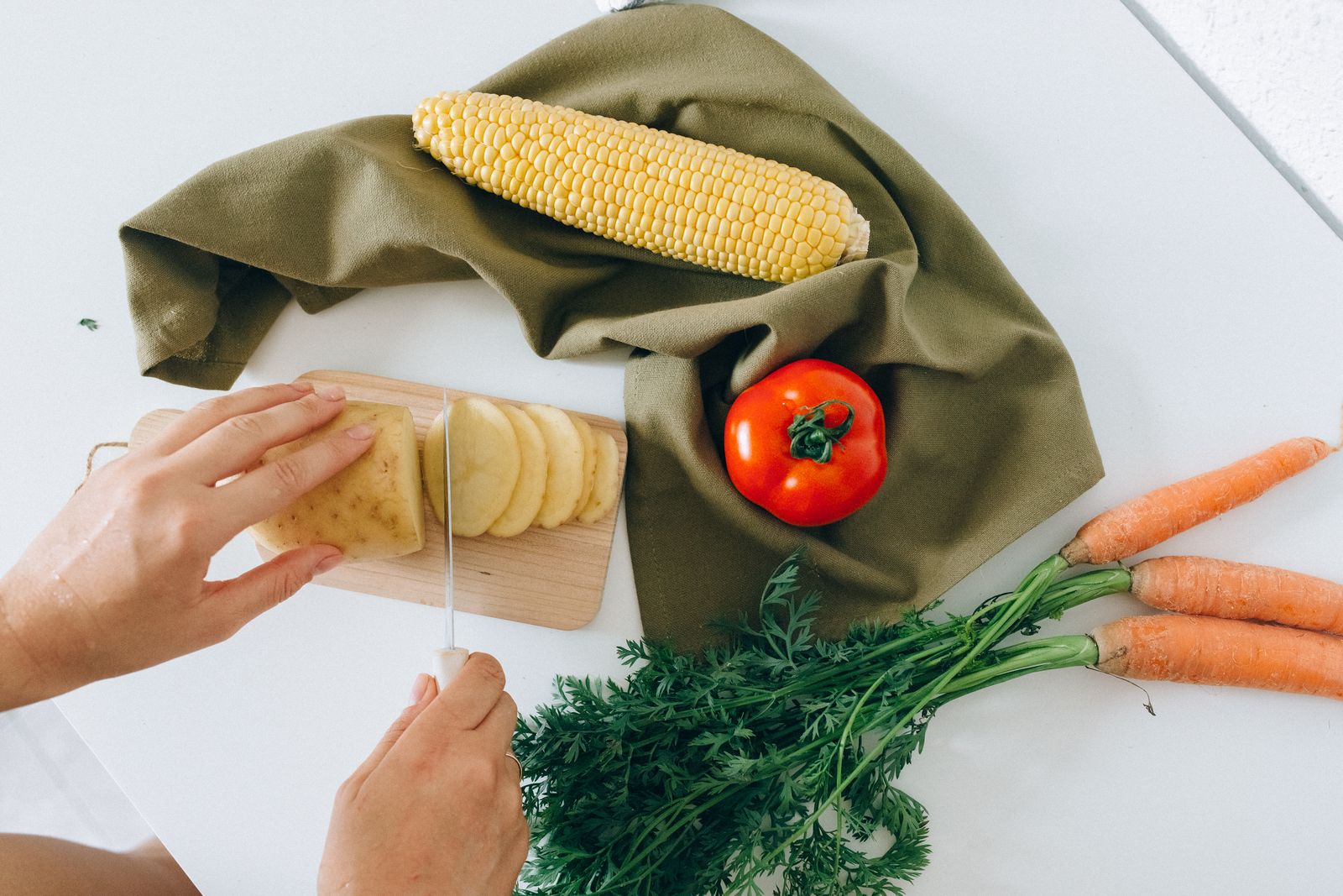 Hands slicing a potato beside vegetables on a preparation surface