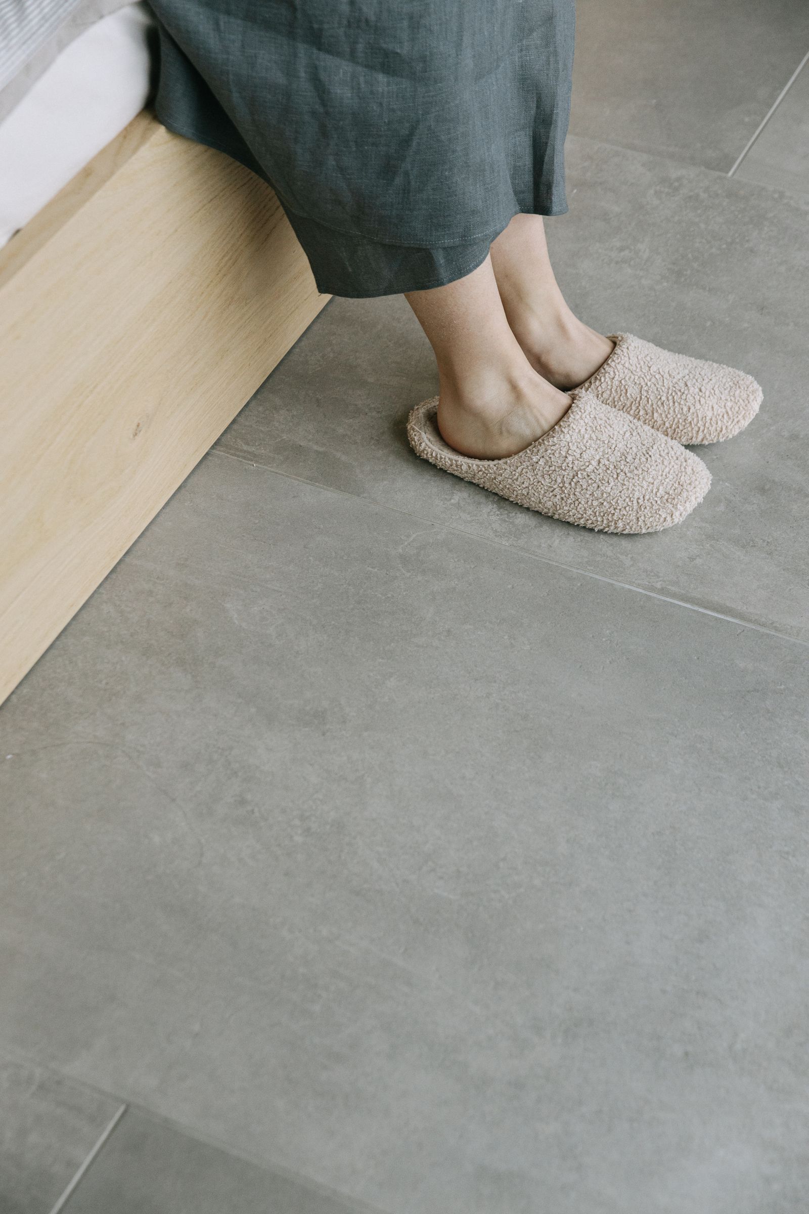 A person wearing beige indoor slippers beside a bed